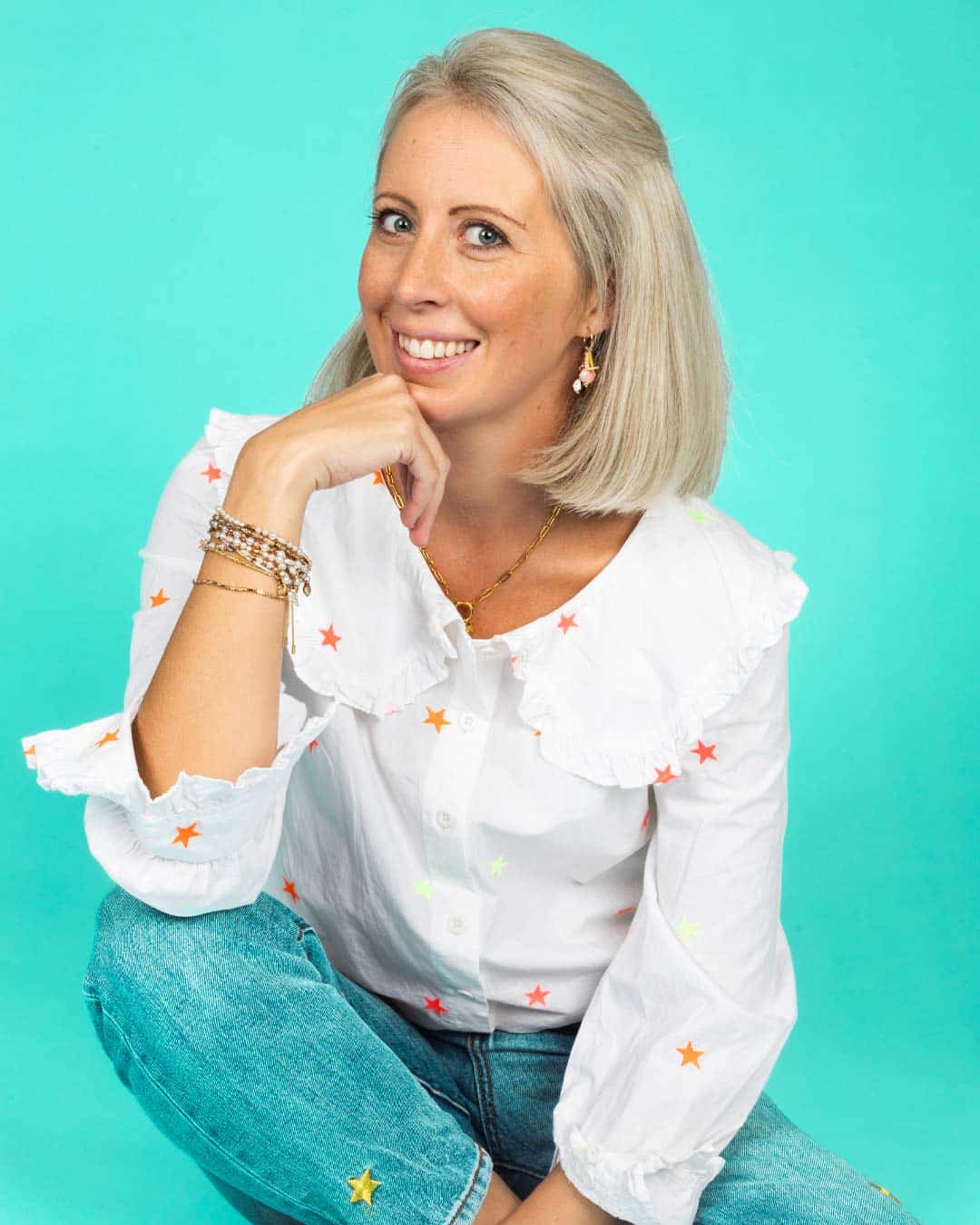 Smiling woman sitting against a turquoise background, wearing a white star‑patterned blouse and jeans.