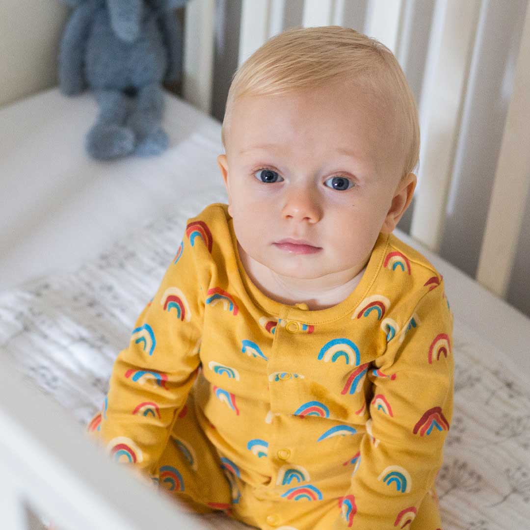 Baby sitting in a crib wearing a yellow rainbow onesie, looking up calmly in a bright nursery setting.